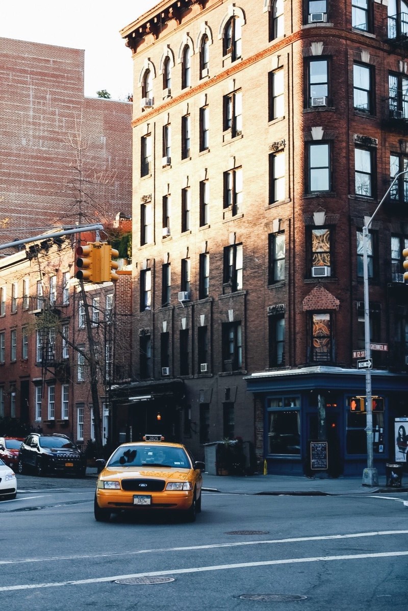yellow taxi on a street in front of a sunny brick building with a streetlamp, yellow cab intersection 4k