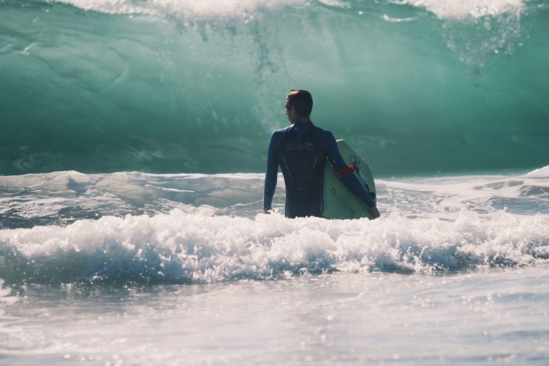 a man in a wetsuit stood in the sea holding his board looking out at a massive onrushing wave, surfer watching the onrushing wave 4k