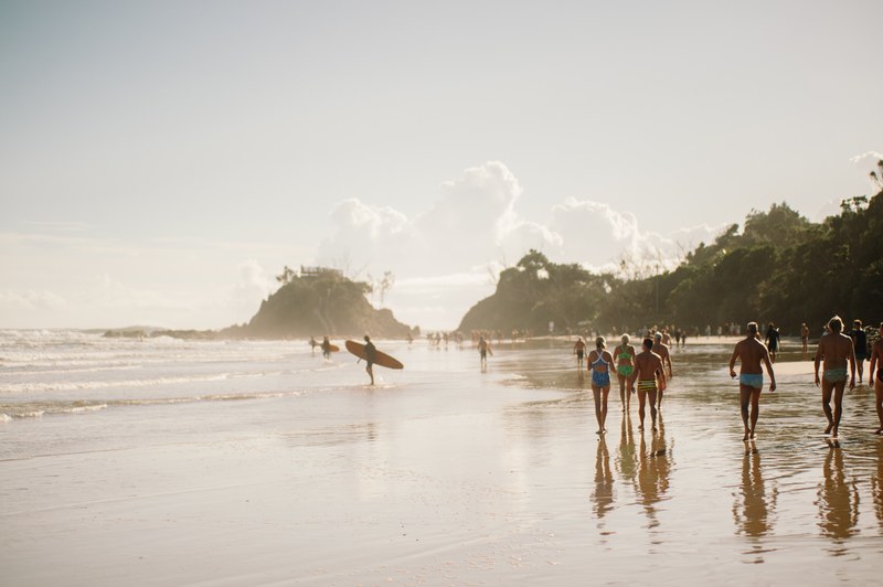 beach goers walking down the tree lined shore as surfers carry their boards into the sea at byron bay, group at byron bay beach 4k