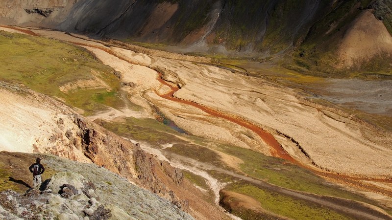 a man with a backpack standing on a rocky ledge looking off into an arid riverbed below, red river below 4k