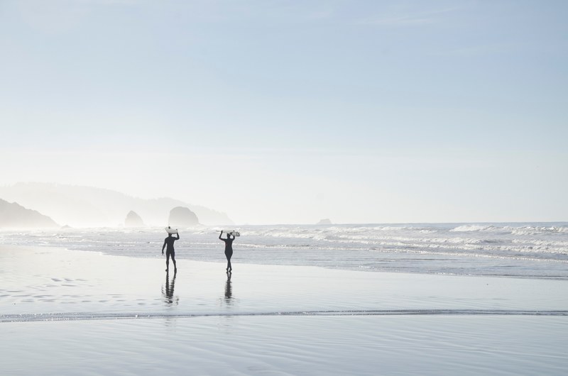 silhouettes of two people carrying surfboards above their heads on a wet beach, surfer silhouettes on cannon beach 4k