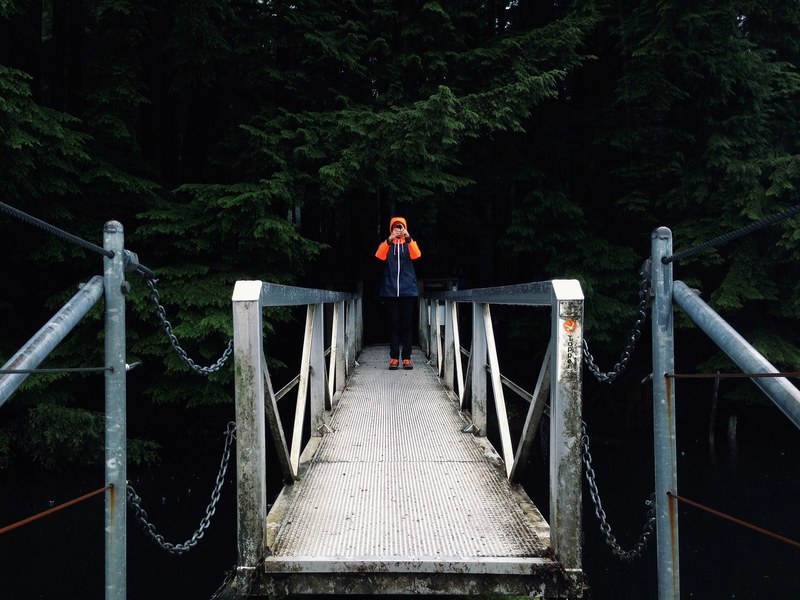 a man on a bridge leading into a forest taking a photo with his phone, taking photos on a bridge 4k