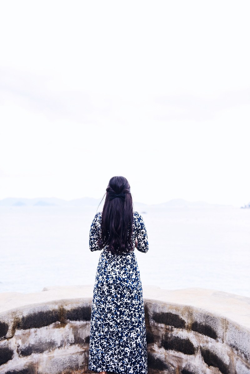 a woman in a dress standing steading next to a stone ledge and looking off into the sea, woman looking off into the sea 4k