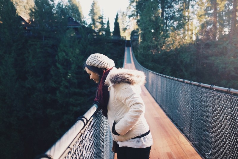 a woman in a knit cap looks over the chain link fence on the capilano suspension bridge, woman on suspension bridge 4k