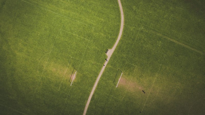 a drone shot of a narrow path with green football fields with goalposts on both sides, drone view of dirt road between soccer fields 4k