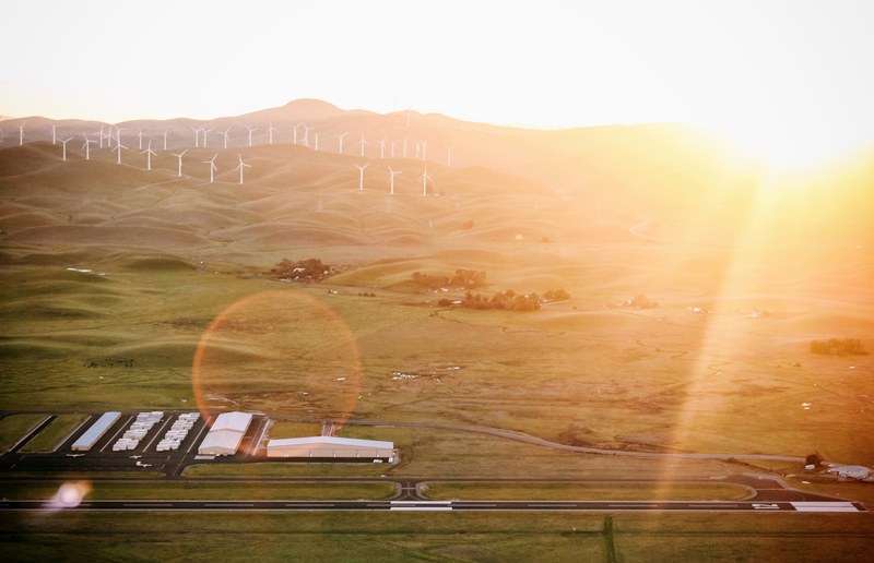 a drone view of byron airport the runway and hangers as well as the surrounding countryside including grassland hills and a wind farm with sun white glare of sunrise or sunset in the distanc 4k