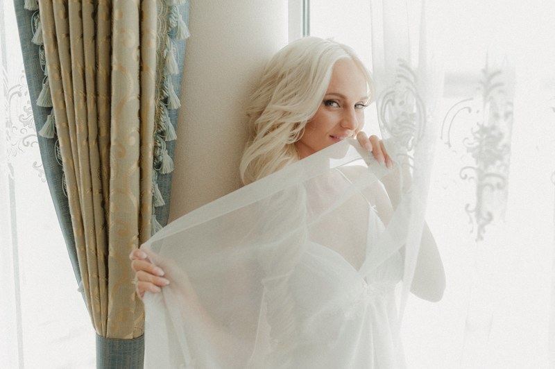 a smiling blonde woman in white gauzy lingerie poses by a window and curtain, woman in white lingerie 4k