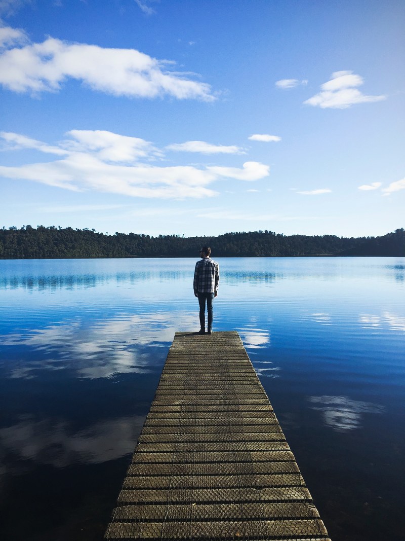 a man wearing a flannel standing on the edge of a dock on a lake that is reflecting the blue sky and clouds, on the piers edge 4k