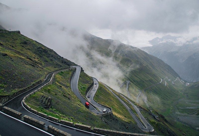 cars driving down a windy road down a mountain covered in mist, stelvio pass 4k