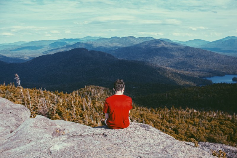 a man sitting on a rocky ledge overlooking wooded hills, ledge over the hills 4k