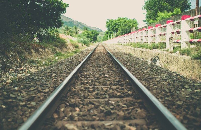 a perspective shot of a railroad track running down a path beside a red and white striped fence several trees and a mountain, _countryside railway 4k