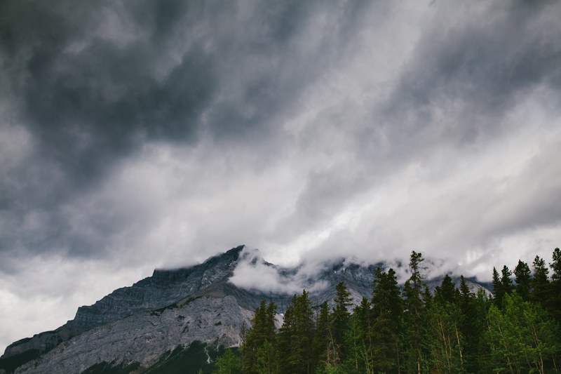 clouds envelop a rugged mountain with a coniferous forest at its base, clouds devouring a mountain 4k