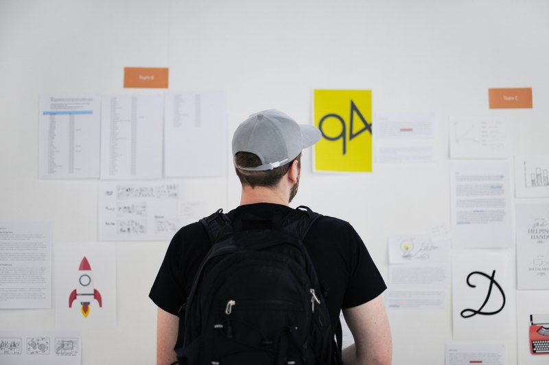a man wearing a black backpack and baseball cap staring at work related documents on the office wall, checking workplace progress 4k