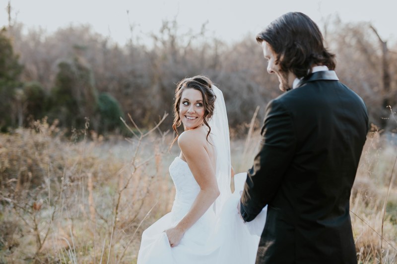 couple on their wedding day in a grassy field the bride smiling back toward the groom, groom follows behind bride 4k