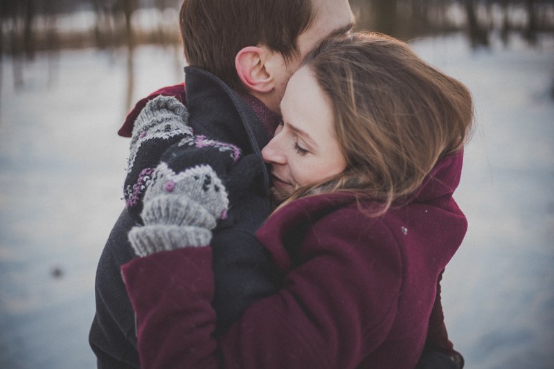 couple in winter clothes hugs outdoors with snow on the ground and trees in the background, hugging in the winter 4k