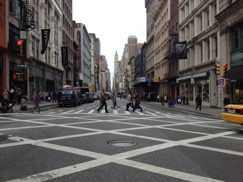 people walking across city street with clothing stores parked cars and skyscrapers in the background, people crossing street 4k