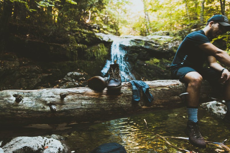 a hiker sitting on a fallen tree stump next to a pair of hiking boots in front of a waterfall, hiker resting on a log 4k
