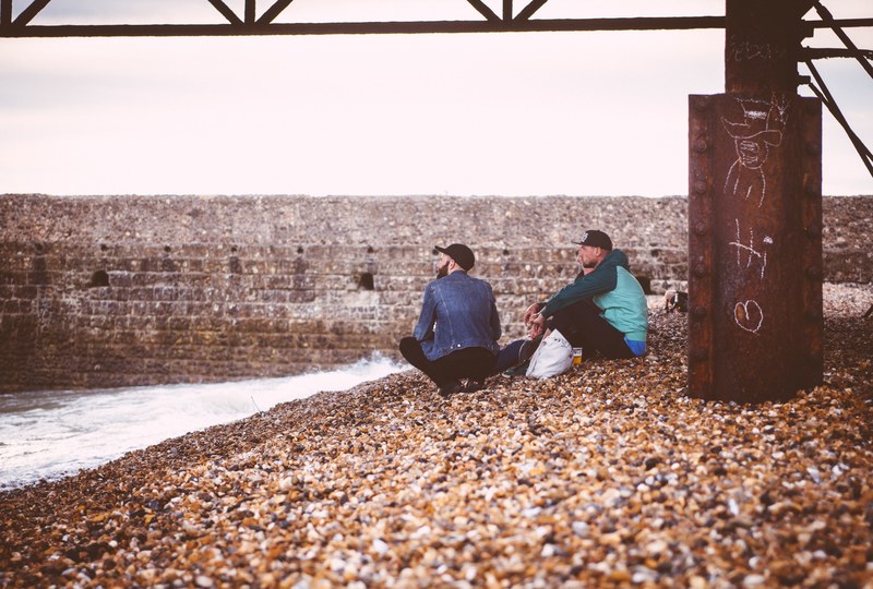 couple of men sitting on the rock pebble coast underneath a pier construction in brighton, men sitting on a pebble beach 4k