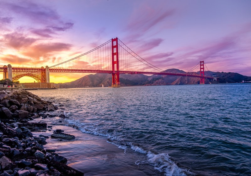 a purple and yellow sunset behind golden gate bridge in san fransisco with waves breaking on the shoreline, golden gate purple sunset_ 4k
