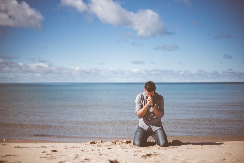 a man in a shirt and jeans kneeling on the edge of a beach with his hands clasped against his chin, gesture of humility 4k