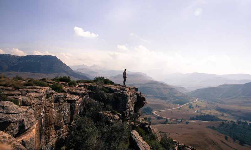 a man standing on the edge of a cliff overlooking a winding road in sani pass, sani pass cliff 4k