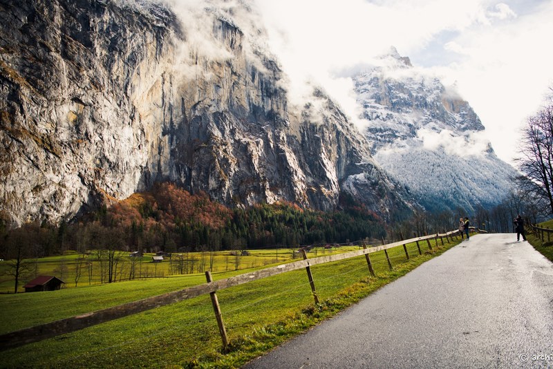 a couple taking their photos with a photographer on a long road with snow covered mountains in the background and a lush green forest in switzerland, _couple photoshoot in switzerland fores 4k