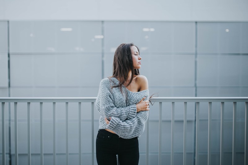 a woman in an off the shoulder sweater stands in front of a white railing and white wall, woman by white railing 4k