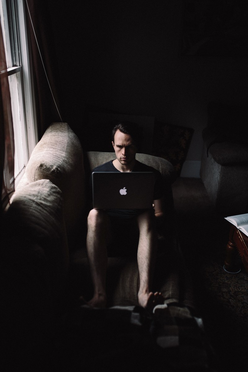 a barefoot man sitting on a sofa and looking at a macbook on his lap, working from a sofa 4k