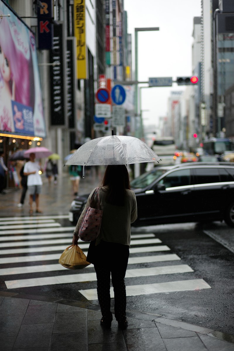 a person holding a clear umbrella waits on the sidewalk to cross a city street, waiting to cross the street 4k