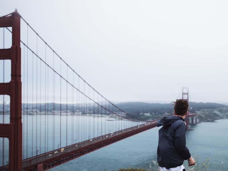 a man in a jacket standing in an elevated spot overlooking the golden gate bridge, golden gate bridge on a windy day 4k