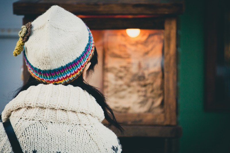 back of a woman with black hair wearing a white jumper and a knit hat staring at a parchment of paper lit and encased in glass, woman in a museum 4k