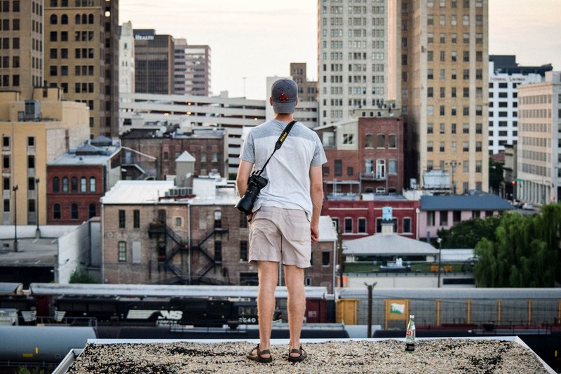 a person in a backwards cap with a camera stands on a rooftop overlooking a city, person with camera on roof 4k