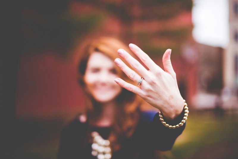 a blurry woman in the background holds up the engagement ring on her hand, engagement ring display 4k