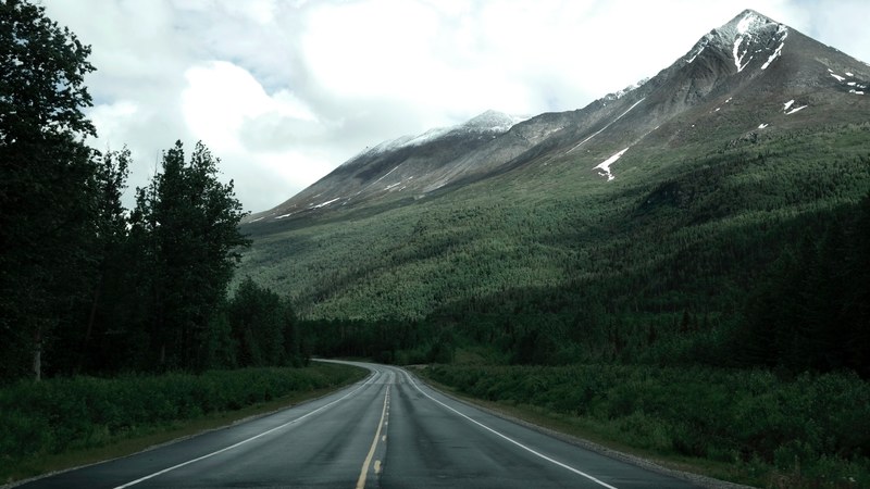 an empty asphalt road near a wooded slope of tall snow capped mountains, road along alaska mountains 4k