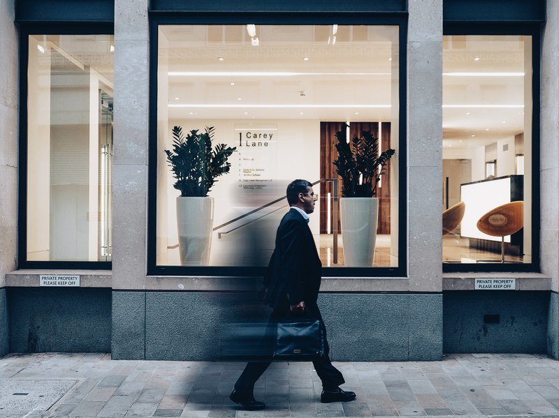 a man in a suit with a suitcase walking past an office building in london, long day in the city 4k