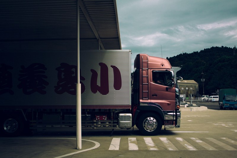 a pink truck with japanese lettering on its container exiting a roofed area beside a hill of trees, pink japanese truck 4k