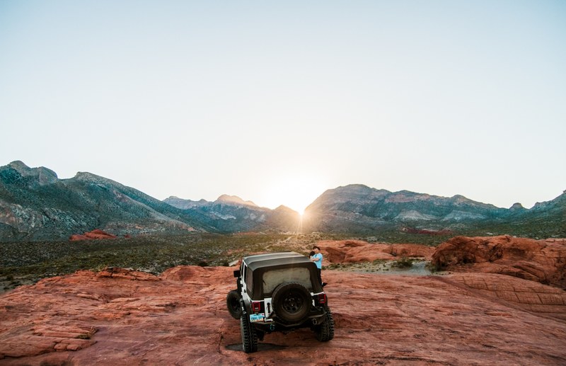 a person and a 4x4 jeep at red rock canyon national conservation area with the sun rising or setting through the mountains on the horizon, a person and 4x4 in a valley and mountain range 4k