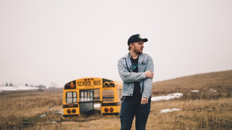 a man wearing a denim jacket holding his arm while staring down the street standing in front of a crashed school bus in a country field, school of wilderness survival 4k
