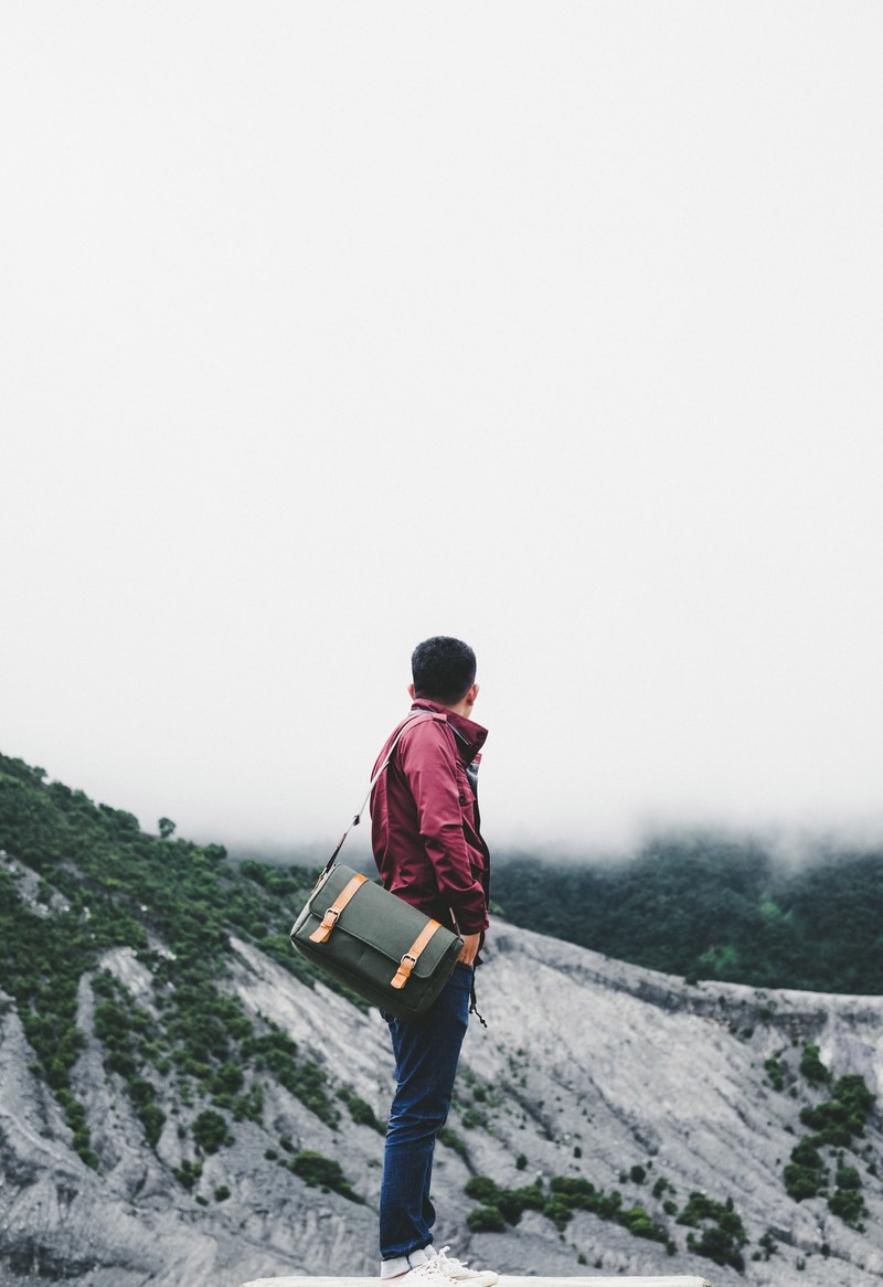 stylish man dressed in jeans and red jacket with leather satchel standing in gray mountain terrain, man red coat mountains 4k