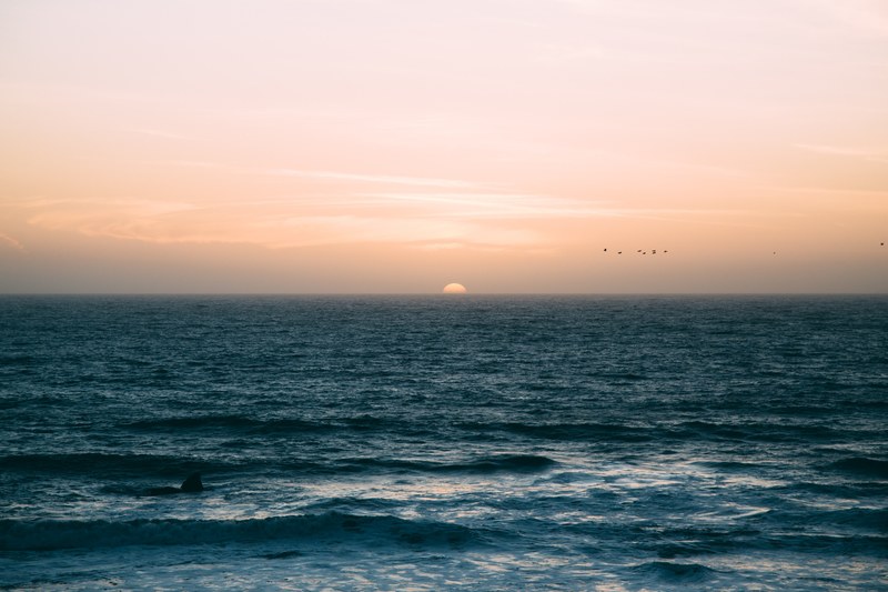 the sea ripples towards half moon bay at the sunrise or sunset lights the sky pale orange and a group of silhouette birds fly past, ocean view towards the pale yellow horizon 4k