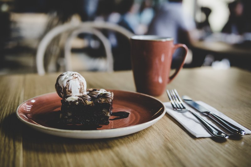 a chocolate nut brownie topped with vanilla ice cream and chocolate syrup beside a mug of coffee and cutlery on a wooden table, delicious bokehlicious 4k