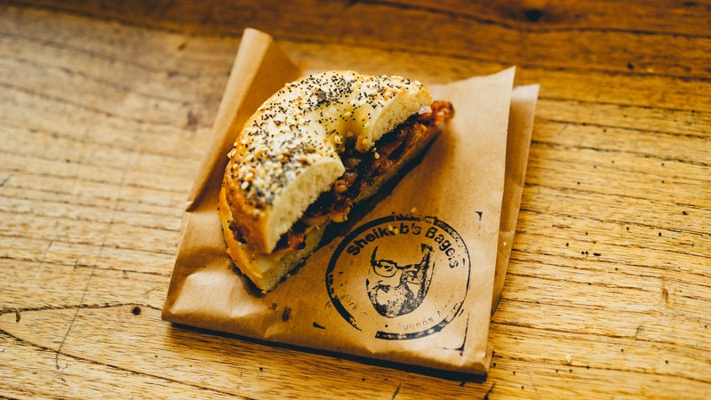 a bacon egg bagel covered in poppy and sesame seeds on top of a brown paper bag labeled shekobs bagels, bacon egg bagel 4k