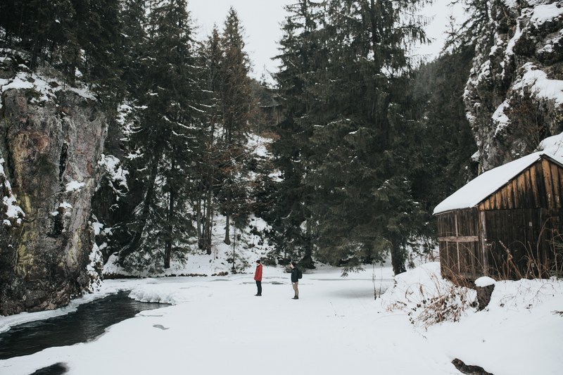 two friends in snowy grda de sus staring at a stream behind a wooden cabin, friends in a snowy forest 4k