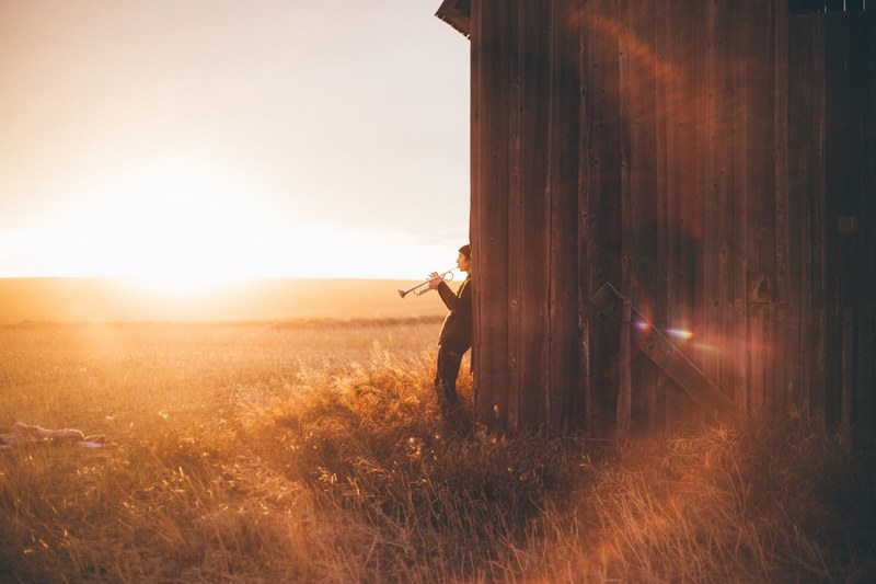 a man plays the trumpet leaning back against a barn wall and facing a golden field, trumpeter in a rural setting 4k