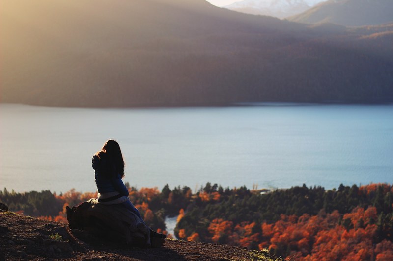 a woman wearing a winter coat sitting on a rock looking at the mountains lake and forest in quilaquina, patagonia view 4k
