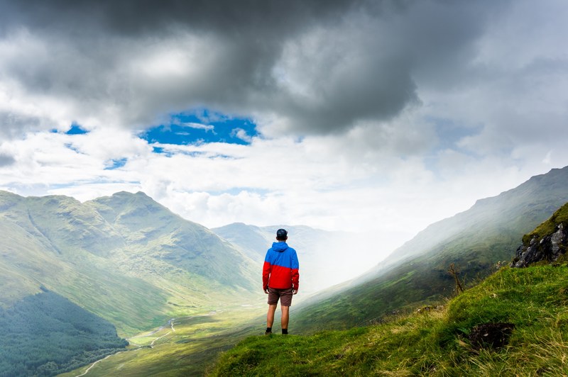 a man wearing a hat a black and red jacket and shorts standing on a grassy mountain in inverlochlarig farm, exploring the mountainside 4k