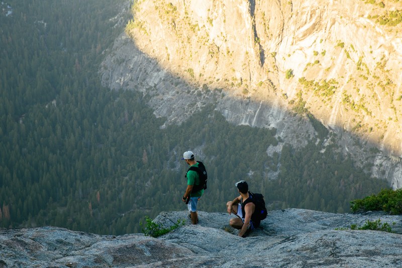two hikers with backpacks resting on a rocky cliff top looking down at vista below, two hikers cliff top 4k