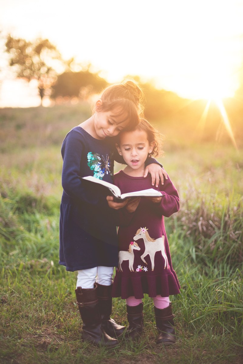 two young girls stand reading from a book outdoors in the sun, girls and book 4k