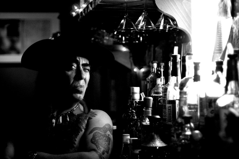 black and white image of a tattooed man sitting next to bottles of alcohol at rite spot cafe, man sitting by alcohol bottles 4k