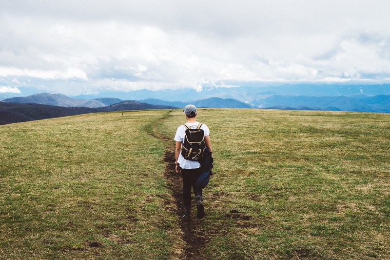 man wearing a hat and backpack hikes away from the camera in green grass toward mountains, hiking man in green field 4k